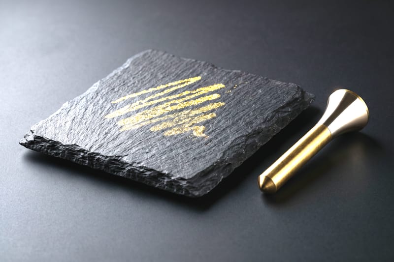 An assayer's black basalt touchstone streaked with gold beside a brass needle.